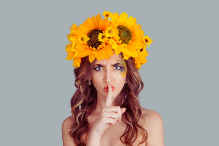 Hush. Woman with floral headband wide eyed asking for silence, secrecy, finger on lips shh hand gesture gray background wall. Fashion girl with crown from sunflowers on head isolated gray background.の写真素材