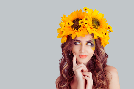 Beautiful thoughtful pensive woman with floral headband looking up daydreaming positive face finger on cheek. Fashion girl with crown from sunflowers on head isolated on gray background with copyspaceの写真素材