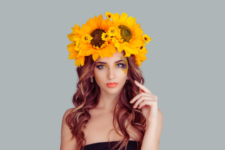 Beautiful young woman wearing floral headband isolated on gray background. Yellow sunflowers wreath of flowers on head looking at you smiling slightly hand touching hair, posing cute gorgeous girl.の写真素材