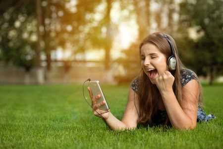 Cute teenager holding smart phone in hand, screams with joy and happiness, surprise with winner expression on face outdoors lying down on green lawn headphones on head. Multicultural model, mixed raceの写真素材
