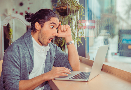 Man shocked looking at laptop. Closeup portrait of a handsome guy wearing formal white shirt and gray blouse sitting near window at a table in living room or coffee shop. Mixed race indian and turkishの写真素材