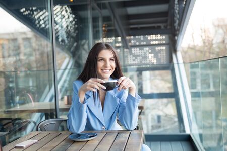 Pensive happy woman remembering looking at side up sitting on a terrace of bar coffee shop restaurant hoping thinking of love, hispanic girl wearing formal blue suitの写真素材