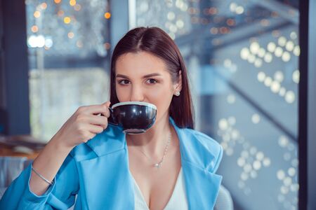 Portrait of smiling pretty woman with cup of coffee. Closeup portrait of beautiful woman hispanic girl wearing formal blue suit sitting at table in a cafe, living room metallic staircase on backgroundの写真素材