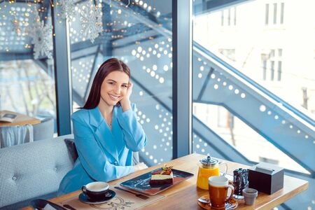 Enthusiastic Woman in trendy autumn outfit chilling in cozy cafe, adjusting hair, happy emotions. Traveling woman. Hispanic girl wearing formal blue suit sitting at a table on a cafe terrace balconyの写真素材
