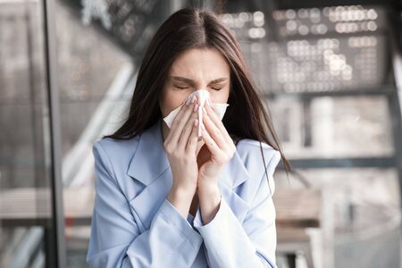 Young woman got nose allergy, flu sneezing nose. Closeup portrait of a beautiful hispanic girl wearing formal blue suit sitting at a table on a cafe terrace balcony outside staircase backgroundの写真素材