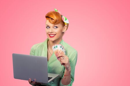 On-line job, internet lottery winner. Closeup red head beautiful young woman girl daydreaming smiling pinup girl green button shirt holding pc, euro bills looking at you camera retro hairstyle on pinkの写真素材