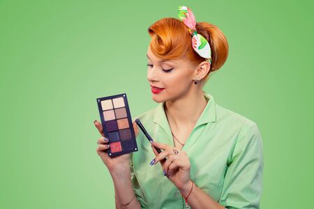 Creative beauty makeup artist. Closeup red head beautiful young woman smiling pinup girl green button shirt holding eye shadow palette brush looking at it, over green background, retro vintage styleの写真素材