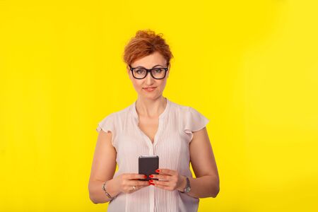 Closeup portrait of a beautiful woman holding using smart phone looking at you camera, girl wearing nude color shirt with retro hair style up, cutout isolated on a yellow backgroundの写真素材