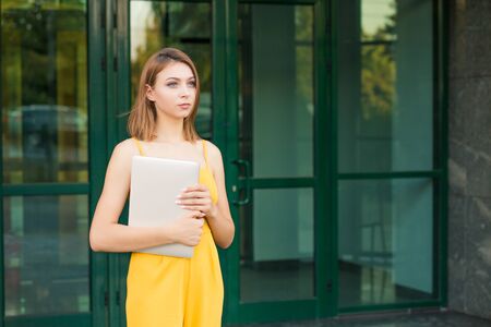 Portrait of pretty woman holding computer laptop posing looking to side thoughtful. Girl standing outdoors near a green building made of glass in a yellow jumpsuit with long bob haircut.の写真素材