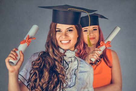 Portrait closeup beautiful happy  graduates, two graduated student girls, young women in cap gown turning smiling holding diploma scroll isolated gray background wall. Celebrating graduation ceremonyの写真素材