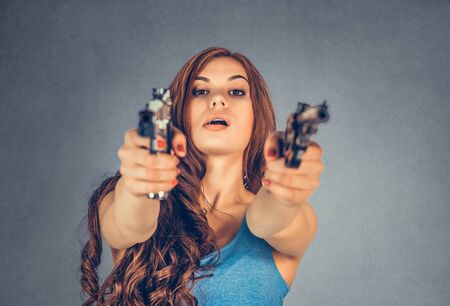 Beautiful young woman with 2 guns. Brunette holding Handguns looking, aimed, pointing at camera. Violent rebel gangster bandit self-justice concept. Mixed race model isolated on gray, grey backgroundの写真素材