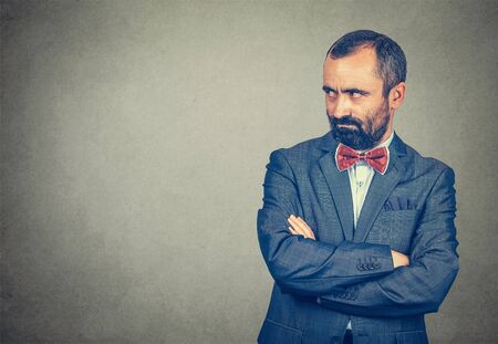 Jealousy, envious. Serious adult gloomy bearded man in elegant suit with red bow tie on gray background with his arms crossed looking envious to side, sideways to side. Negative emotion, body languageの写真素材