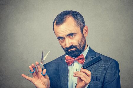 Bearded hairdresser, wearing elegant jacket with red bow tie, posing with scissors and comb. Stylish adult man studio portrait, isolated at grey background. Human face expressionの写真素材