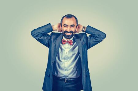 Happy, large smiling businessman keeping hands behind his head looking surprised at you camera. Human gestures. Happy man with dark suit, blue shirt, red bow, tie. Uniform green yellow background.の写真素材