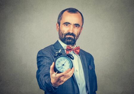 Portrait of a businessman office manager, man in formal clothing holding showing alarm clock, deadline concept. Mixed race bearded model isolated on gray background with copy space. Horizontal image.の写真素材