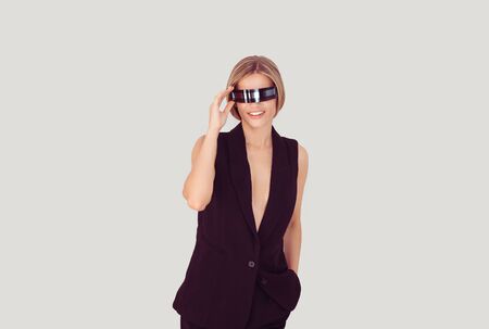 Smiling robot female concept. Beautiful woman putting touching futuristic robot glasses with one hand on the head holding another hand in pocket. Posing over light gray white backgroundの写真素材