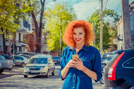 Taxi app. Pretty happy smiling urban woman using smart phone, standing on city street on sunny day waiting for taxi. Beautiful redhead girl holding mobile, texting sms message wearing jeans shirtの写真素材