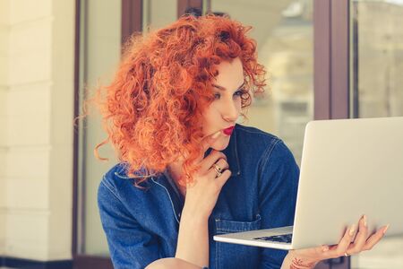 Young woman holding notebook, looking skeptical to screen, touching chin with hand, sitting outdoor....