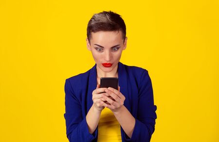 Stunned by sms. Portrait surprised woman typing on smart phone by news saw on cell mobile phone. Multicultural Latina girl short hair red lips blue suit yellow shirt isolated yellow background wallの写真素材