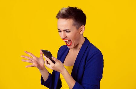 Portrait angry young woman screaming looking on mobile phone isolated on yellow wall background. Negative human emotions feelings. Short hair, blue suit, yellow shirtの写真素材