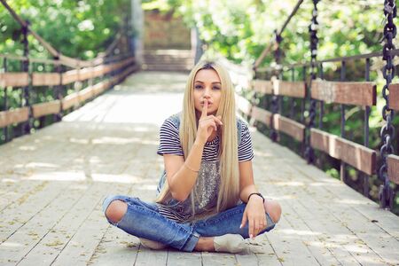 Attractive blonde in stylish summer outfit sitting on bridge in park touching lips with forefinger asking for silence and looking at camera.の写真素材
