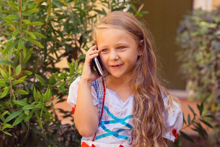 Phone talk happy. Little girl talking on the phone, excited and happy explaining something to her mother nanny father parents isolated green shrubs nature near house outdoors on the backgroundの写真素材