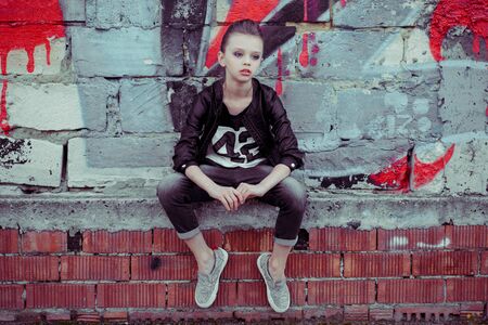 Little fashionista. Closeup portrait head shot beautiful blonde young woman with trendy clothing style isolated sitting on graffiti background wall looking to the side. Neutral face expressionの写真素材