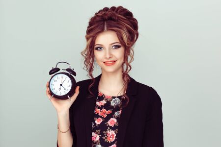 Portrait young funny looking smiling business woman holding alarm clock waiting isolated light green wall background. Human face expression emotion Time punctuality busy schedule conceptの写真素材