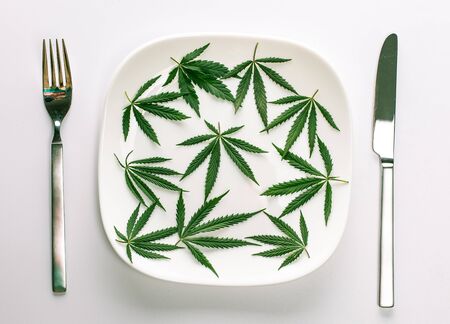 Conceptual image of a green cannabis leaves on a white plate with a knife and fork isolated on a white background with copy space for text. Don't take drugs, take natural herbal herbs medicine conceptの写真素材