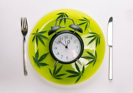 Time for Weed Pot Cannabis Clock Deadline Concept. Alarm clock Marijuana leaves around it on a light green plate with fork and knife Cutlery isolated on white gray backgroundの写真素材