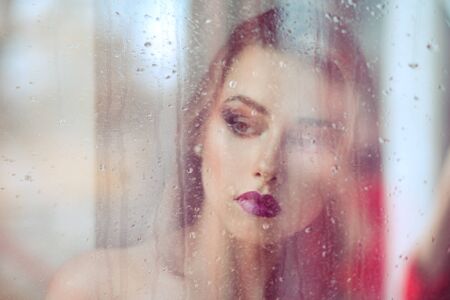A beautiful melancholic alone sad woman stands in front of the window, looking down thinking in...