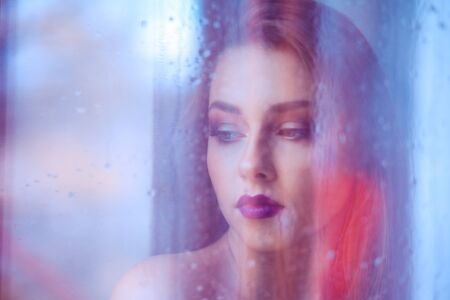 A beautiful melancholic woman stands in front of the window, looking down thinking. Image made through the window, rain on window and reflection on outdoors scene.の写真素材