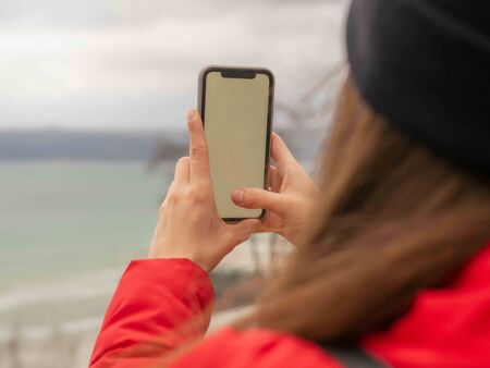 Woman in red coat and black cap holding a cellphone with blank screen standing outside taking a...