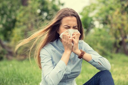 Brunette girl woman sneezing in a tissue blowing her runny nose outdoors, outside backgroundの写真素材