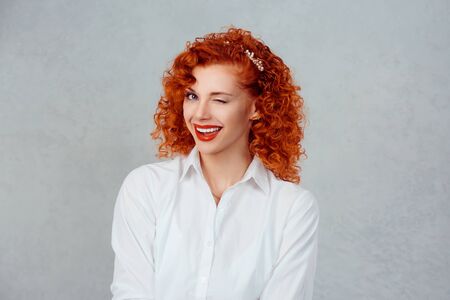 Successful business woman. Closeup portrait head shot sexy pretty confident redhead curly girl winking isolated gray background wall. Positive human emotion face expression feeling life perceptionの写真素材