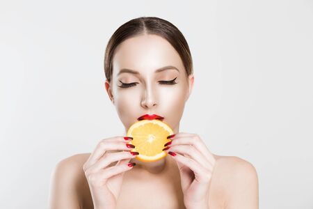 Great food for a healthy lifestyle. Beautiful young shirtless woman holding  having a slice piece of orange in her mouth while standing against white grey backgroundの写真素材