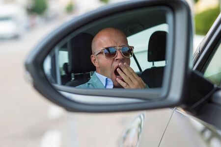 Side mirror view reflection sleepy tired fatigued yawning exhausted young man driving his car in...
