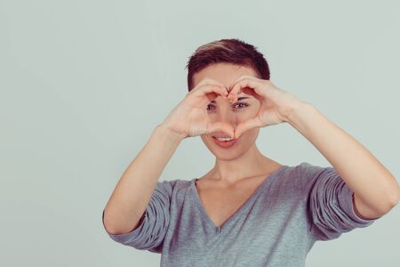 Happy beautiful young woman shows heart shape hands on bright green grey background. Positive human emotions, face expressions, feelings, body language, life perceptionの写真素材