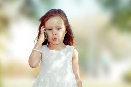 Kids & technology concept. Closeup portrait headshot beautiful upset serious little girl  talking mobile having conversation green park outdoor background. Multicultural mixt race asian russian modelの写真素材