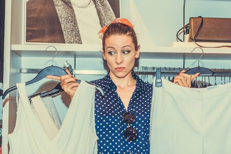 Pretty woman feeling uncertain about what to buy choosing deciding between two outfits on hangers in...