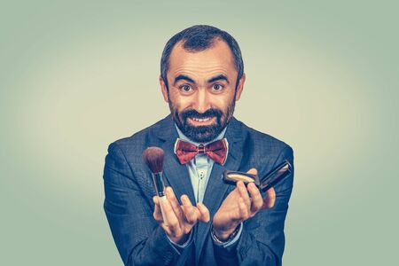 Funny business man holding brush and compact loose powder holding a mirror looking at you camera showing the makeup kit. Mixed race bearded model isolated on green yellow uniform gradient backgroundの写真素材