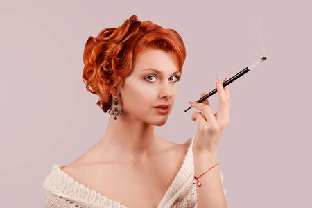 A beautiful young woman retro beauty girl up hairdo style posing in studio smoking a cigarette while standing in front of a pink background. Fashionable white wool dress natural make-up and tan skinの写真素材