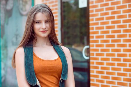 Woman smiling outdoors. Closeup portrait woman girl looking at you camera smile on face orange copper shirt blue jeans jacket looking at you camera outside her home brown bricks wall grunge backgroundの写真素材