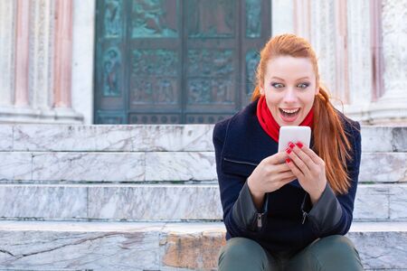 Shocking SMS. Closeup portrait funny shocked young girl looking at phone receiving good news photos message with stunned emotion on face sitting on the stairs of a city. Model in blue coat red scarfの写真素材