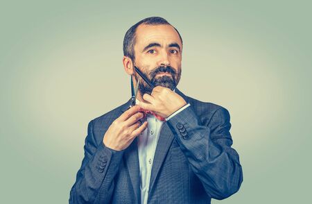 Bearded hairdresser, barber styllist cutting styling his own beard with scissors and comb, wearing elegant jacket with red bow tie. Stylish adult man studio portrait, isolated at green backgroundの写真素材