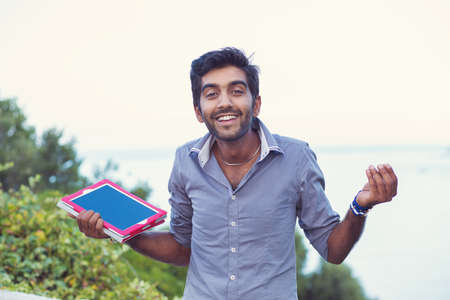 Confused student. Closeup portrait puzzled clueless young man holding, showing tablet pad screen, arm out asking what is problem who cares so what I donât know isolated sea and nature on backgroundの写真素材