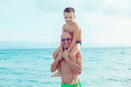 Father giving son ride on back in water. Portrait of happy father giving son piggyback ride on his shoulders and looking up. Cute boy with dad playing outdoorsの写真素材