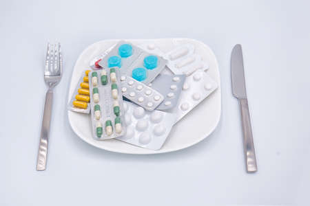 Diet.  Different  pills, medicines, drugs on a white square shaped plate with fork and knife around isolated on white background. Taking pills instead of food, unhealthy sick lifestyle conceptの写真素材