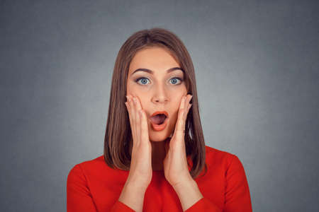 Closeup portrait happy young woman in red looking excited in full disbelief hands on cheeks isolated...