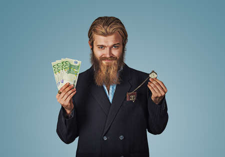 Time is money. Handsome cheerful bearded hipster man smiling with cash and pocket watch. Funny guy is a lucky winner holding a pile of money and a watch he is happy to win the jackpot isolated on blueの写真素材